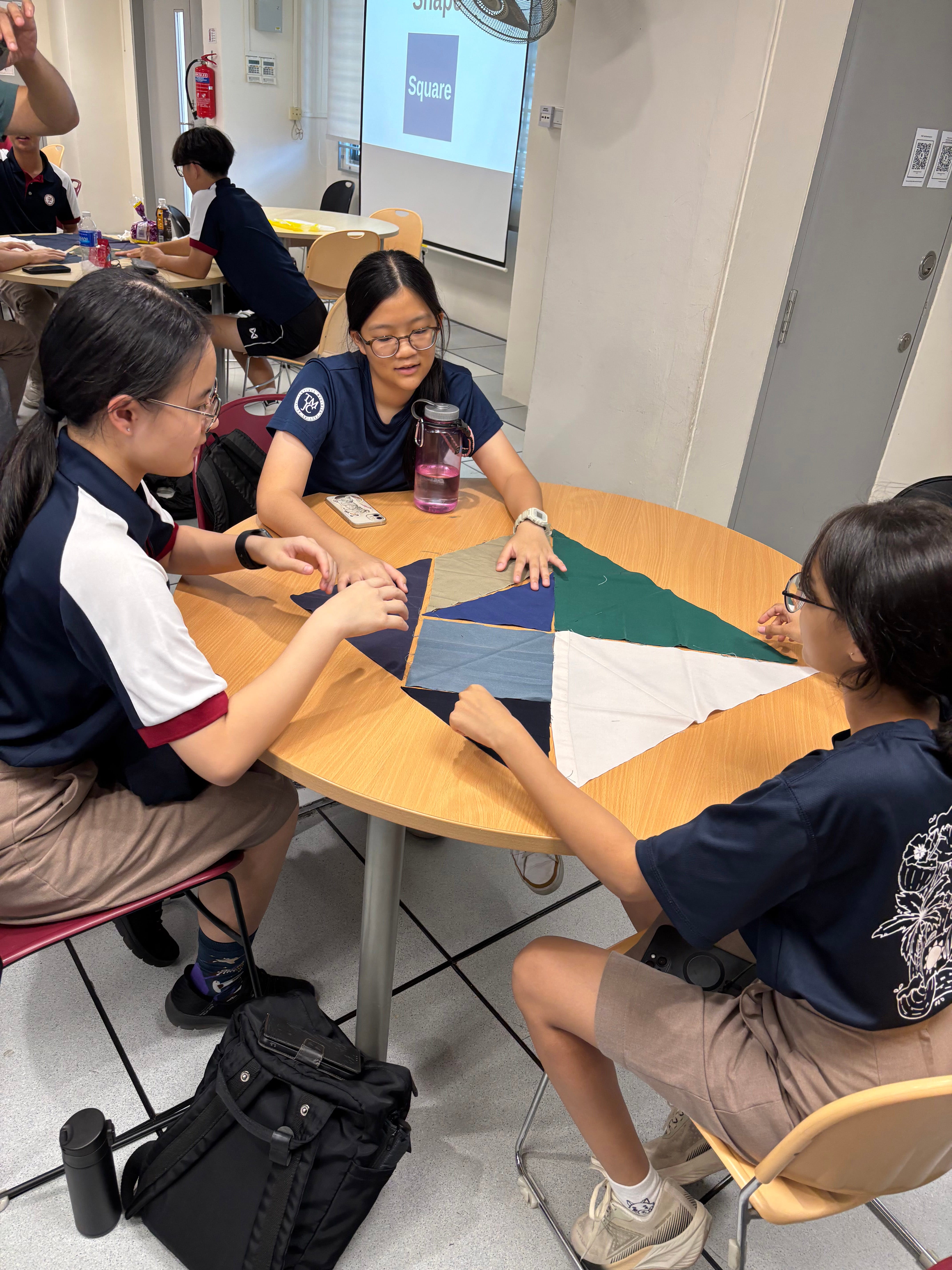 A group of students in navy school uniforms gather around a round table, arranging and examining triangular fabric pieces in navy, green, and white as part of a banner-making activity.